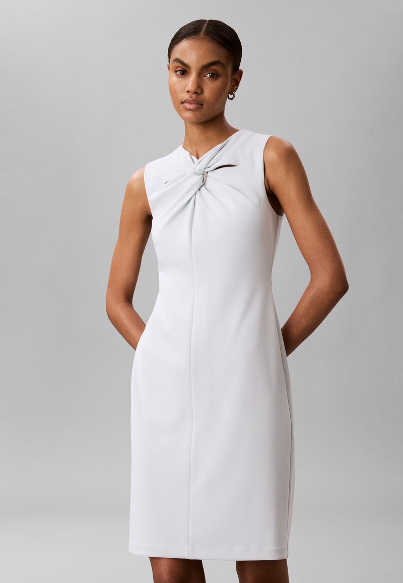 Femme debout, les mains dans le dos, portant une robe blanche sans manches avec un détail de nœud torsadé à l'encolure sur un fond gris.