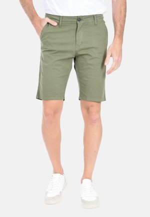 Uomo che indossa pantaloncini verde oliva fino al ginocchio e scarpe da ginnastica bianche, in piedi contro uno sfondo chiaro e semplice.