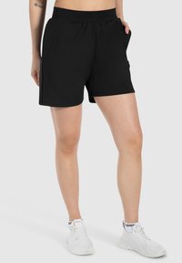 Bermudas deportivas negras hechas de tejido elástico, con una cinturilla elástica, bolsillos laterales y un diseño sencillo. Realzadas por una textura suave.