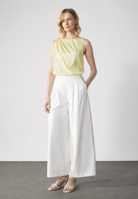 Top giallo senza maniche con dettaglio drappeggiato, abbinato a pantaloni bianchi larghi a vita alta. Design chiaro e minimalista con texture morbide.