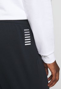 I jogger di un blu scuro presentano strisce riflettenti argentate sui lati, abbinati a una maglietta bianca a maniche lunghe, che mette in mostra un tessuto liscio e una vestibilità rilassata.