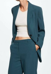 Blazer teal avec des boutons noirs, doté d'un design structuré, associé à un haut cropped côtelé crème et un pantalon tapered assorti.