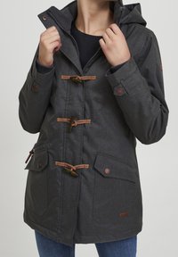 Person som bär en mörkgrå hooded jacka med knäppningar och fickor med klaff, håller kragen nära nacken, ihop med blå jeans.