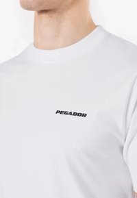 Balta trumpomis rankovėmis marškinėliai, pagaminti iš medvilnės, su juodu "PEGADOR" logotipu kairėje krūtinės pusėje. Lygus paviršius ir klasikinis apvali iškirptė.
