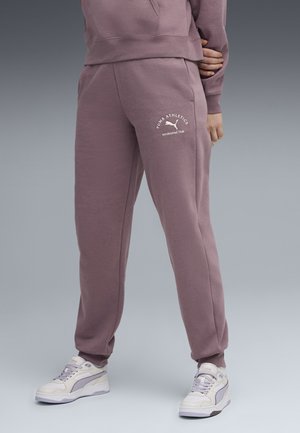 Person trägt mauvefarbene Puma Athletics Jogginghose und passenden Hoodie, kombiniert mit weißen und lila Puma Sneakers, steht mit den Händen hinter dem Rücken.