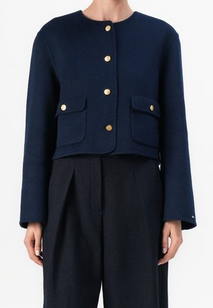 Veste courte bleu marine avec boutons dorés et poches à rabat, portée sur un pantalon plissé noir taille haute.