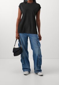 Osoba w czarnej bluzce bez rękawów, luźnych niebieskich dżinsach cargo, w białych trampkach, trzymająca małą czarną torebkę przy boku.