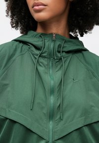 Πράσινο μπουφάν με φερμουάρ και κουκούλα, με υφή, ταιριαστά κορδόνια και μικρό λογότυπο Nike στην αριστερή πλευρά του στήθους.