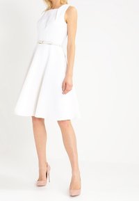 Robe blanche sans manches avec un tissu texturé, jupe évasée et taille ajustée, complétée par une fine ceinture en argent et des escarpins roses clairs.