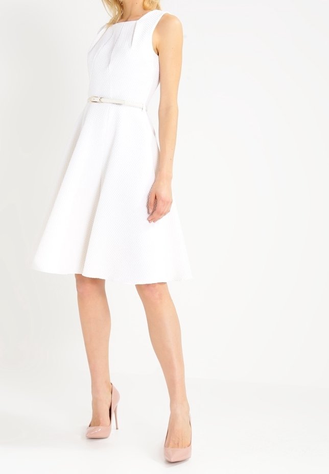 Robe blanche sans manches avec un tissu texturé, jupe évasée et taille ajustée, complétée par une fine ceinture en argent et des escarpins roses clairs.
