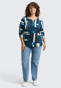 Vrouw met krullend haar, gekleed in een blauw geometrisch patroon met lange mouwen, lichte spijkerbroek en witte sneakers, staande tegen een effen achtergrond.