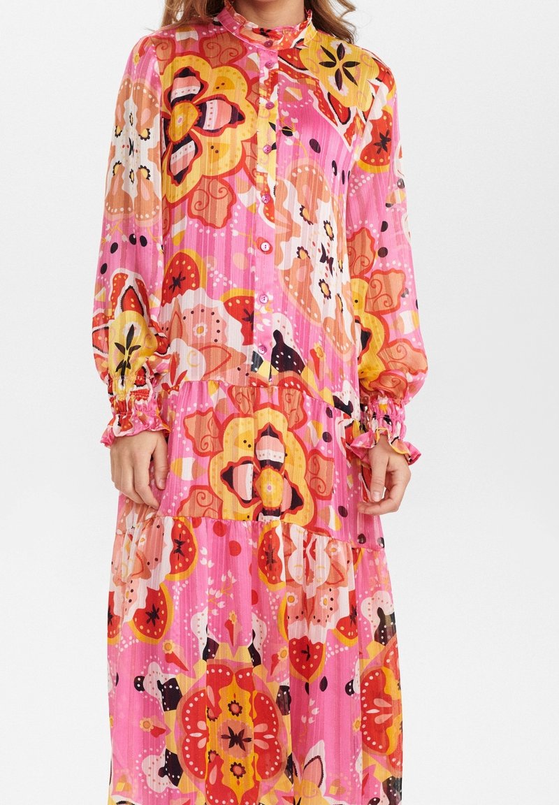 Robe rose à manches longues avec devant boutonné et jupe à volants ornée de grands motifs floraux orange, jaune et rouge.