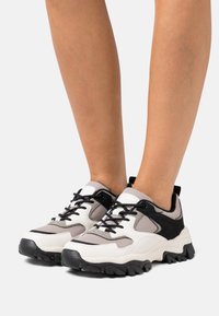 Baskets épaisses blanches et noires avec textures en maille et en daim, dotées d'un accent gris et d'une semelle extérieure noire épaisse. Design à lacets.