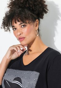 T-shirt nera a collo a V con una grafica geometrica in bianco e nero, caratterizzata da una texture morbida. Il modello ha i capelli ricci e indossa orecchini a goccia.