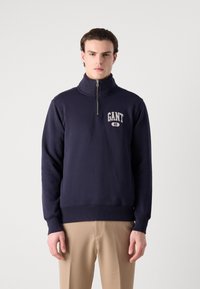 Pulover bleumarin cu guler cu jumătate de fermoar, având un logo GANT. Fabricat dintr-un material moale, cu manșete și tiv cu dungi pentru un fit structurat.