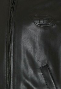 Gros plan sur une veste en cuir noire montrant la fermeture éclair, un logo ovale embossé sur la poitrine et les coutures verticales des poches.