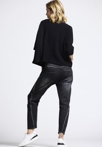 Woman with blonde hair stands sideways wearing a black sweater, faded black jeans, and white sneakers against a plain white background.