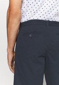Rechterzijde van een persoon in marineblauwe broek met een achterzak met knoop en een wit shirt met kleine blauwe en groene geometrische patronen.
