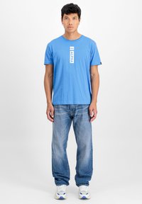 Modra majica s crew neck iz bombaža, ki ima navpični bel logotip 'ALPHA'. Kombinirana z ohlapnimi svetlo modrimi kavbojkami in belimi supergami.