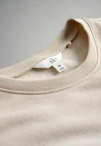 Sudadera beige con cuello redondo acanalado, textura suave y etiqueta de cuidado que indica talla M. Fabricada en Sri Lanka, presenta un diseño minimalista.