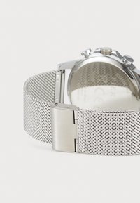 Metallarmbanduhr mit einem silbernen Netzband, rundem Gehäuse und einem Verschluss. Die Rückseite zeigt eine Edelstahlmarkierung und eine glatte, polierte Oberfläche.