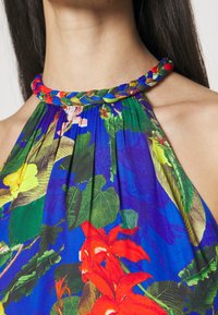 Gros plan sur une robe d'été colorée à encolure dos-nu avec des motifs floraux tropicaux rouges, jaunes et verts, et un détail de col torsadé sur une femme.