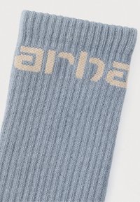 Ανοιχτό μπλε πλεκτό κάλτσα με ριμπ υφή. Διαθέτει μπεζ γράμματα που σχηματίζουν τη λέξη "arha" κοντά στην κορυφή.