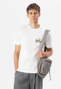 Hvit bomull T-skjorte med "REPLAY" trykt i svart og gull, sammen med en grå stoff crossover-bag som har en frontlomme og justerbar stropp.