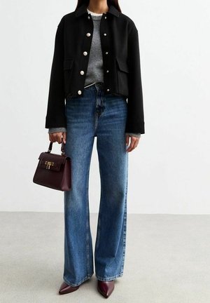 Henkilö, jolla on musta napillinen takki, harmaa villapaita, leveälahkeiset siniset farkut, viininpunaiset kärjestä terävät kengät ja viininpunainen käsilaukku kädessä.