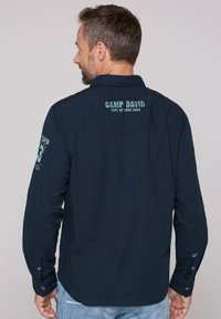 Camicia a maniche lunghe di un blu scuro realizzata in tessuto tessuto, con la scritta ricamata "CAMP DAVID" sulla schiena e sulla manica sinistra. Polsini con bottoni visibili.