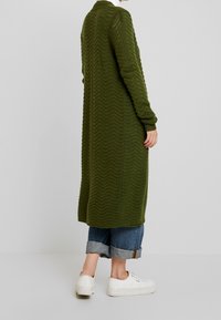 Long gilet vert olive avec un motif texturé en zigzag. Poignets et ourlet côtelés, porté avec un jean bleu retroussé et des baskets blanches.