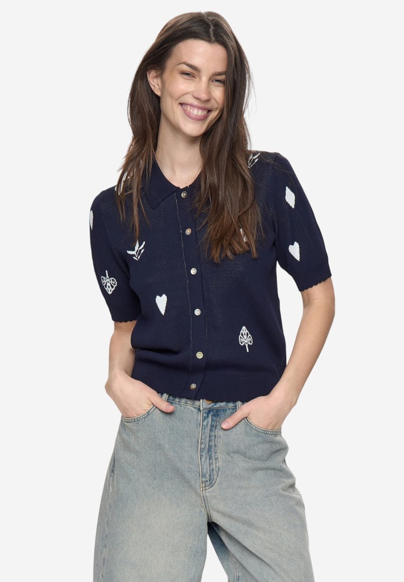 Tmavě modrý kardigan s krátkými rukávy a zapínáním na knoflíky, zdobený bílou výšivkou se vzory srdcí, kosočtverců a listů. Pletená textura.