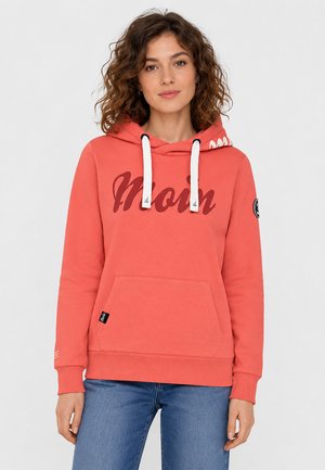 Vrouw met krullend haar die een koraalkleurige hoodie draagt met witte trekkoorden en "Moin" aan de voorkant, gecombineerd met een blauwe spijkerbroek.