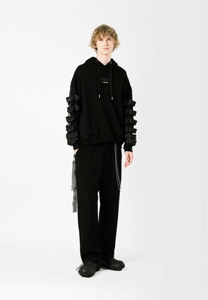 Sudadera negra con mangas en 3D en capas, que presenta un logo central. Combinada con pantalones negros y calzado texturizado. Accesorio de cadena en los pantalones.