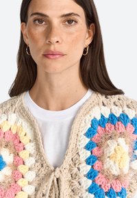 Crocheted vest in cream, pink, blue, and yellow with a floral pattern. Features a tie closure and a textured, plush appearance.