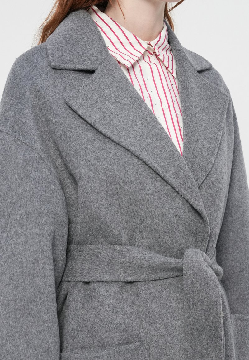 Donna che indossa un cappotto grigio in lana con cintura sopra una camicia bianca a righe verticali rosse e piccoli punti neri.