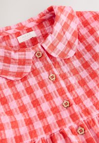 Vestito a quadretti rossi e rosa con bottoni di legno a forma di fiore e colletto arrotondato, etichettato "NEXT Made in India".