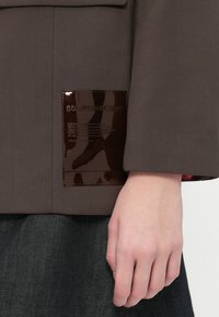 Jachetă maro cu o etichetă lucioasă de culoare maro pe mânecă, având text embosat. Materialul pare neted, cu un design structurat.