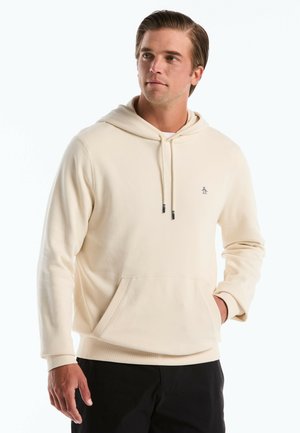 Junger Mann trägt einen cremefarbenen Hoodie mit Fronttasche und Kordelzug, steht mit einer Hand in der Tasche und blickt leicht nach rechts.