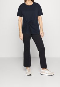 Žena stojí oblečená v námořnickém uzlu t-shirt, černých kalhotách a béžových teniskách, na pozadí jednoduchého bílého pozadí.