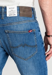 Tył niebieskich spodni Mustang Oregon Tapered z brązową naszywką marki, biała metka wskazująca rozmiar W31 L34, noszonych przez osobę w białej koszuli.