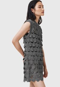 Rochie scurtă, fără mâneci, dintr-un material texturat de culoare neagră, cu detalii în formă de arce și modele circulare. Prezintă un decolteu rotund și o siluetă ajustată.