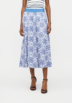 Vrouw draagt een midi-rok met blauwe en witte bloemendessin, een blauwe tailleband en zwarte sandalen met hoge hak en bandjes.