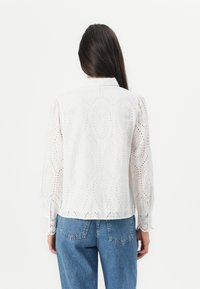 Kvinna med långt mörkt hår bär en vit ögla-blus med lång ärm och blå denimjeans, vänd bort mot vit bakgrund.