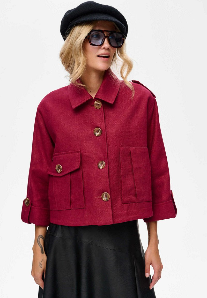 Bordeaux kleurige cropped jas met grote voorzakken, knoopsluiting en een kraag. Voorzien van opgerolde mouwen en dubbele knoopaccenten.