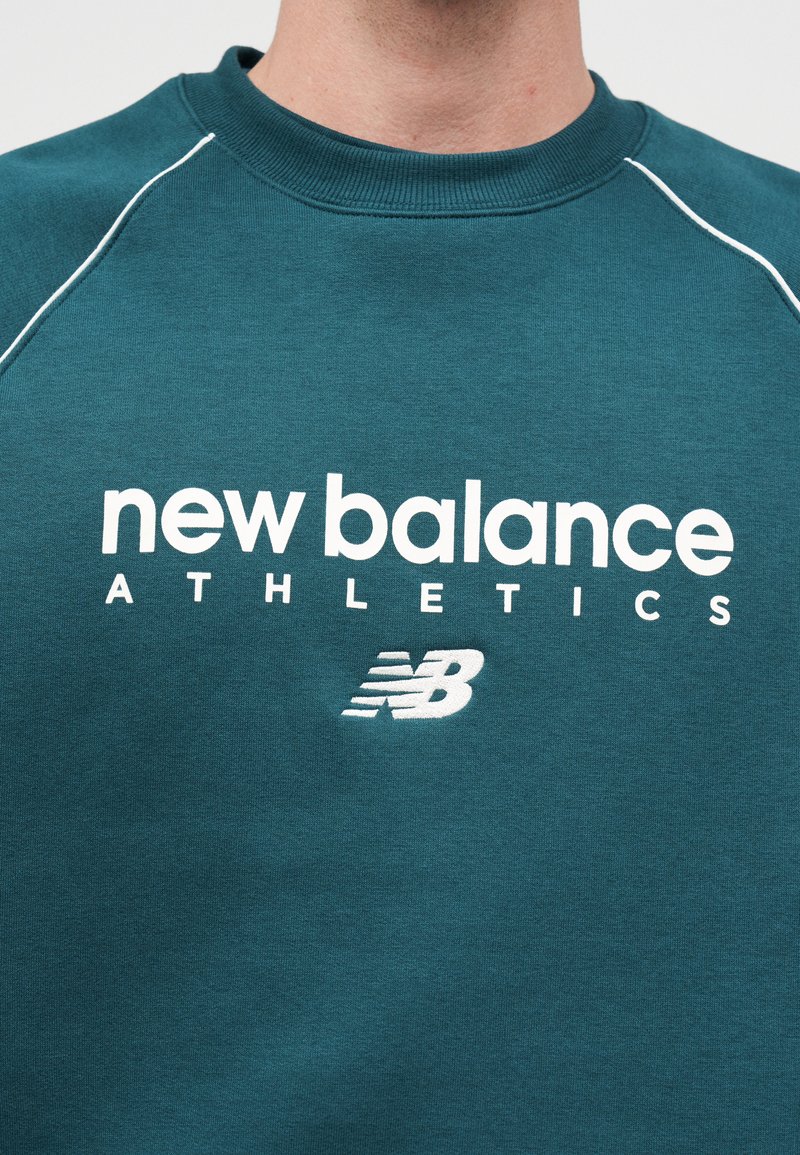 Sweat-shirt bleu sarcelle avec texte blanc "new balance ATHLETICS" et logo "NB" sur la poitrine, porté par une personne visible du cou jusqu'au milieu du torse.