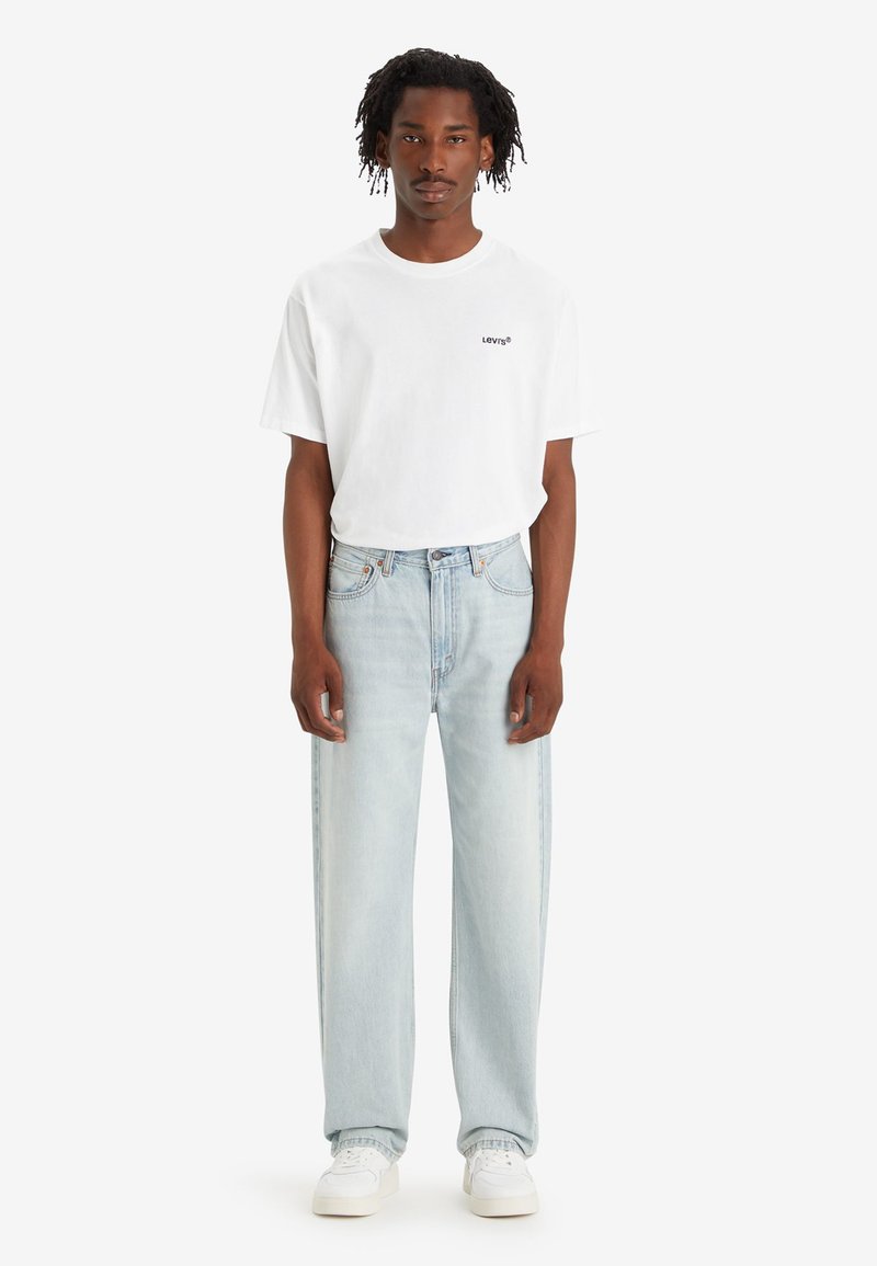 Camiseta blanca de algodón con el logo "Levi's", combinada con unos vaqueros de denim azul claro. El modelo lleva zapatillas blancas y está de pie frente a un fondo neutro.