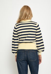 Prúžkovaný pulóver v žltej a námornej farbe, s vysokým golierom a balónovými rukávmi. V kombinácii s tmavými džínsami.