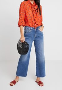 Blusa floral roja con lazo, jeans ajustados de talle alto azules, bolso estructurado negro y sandalias rojas de dedo.