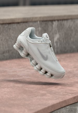 Hvide Nike-sko med en glat, struktureret overdel og lynlåslukning, med en buet sål med synlige polstringspuder.